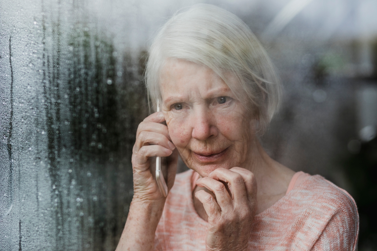 Senior woman is looking worriedly out of the window of her home while talking to someone on the phone.